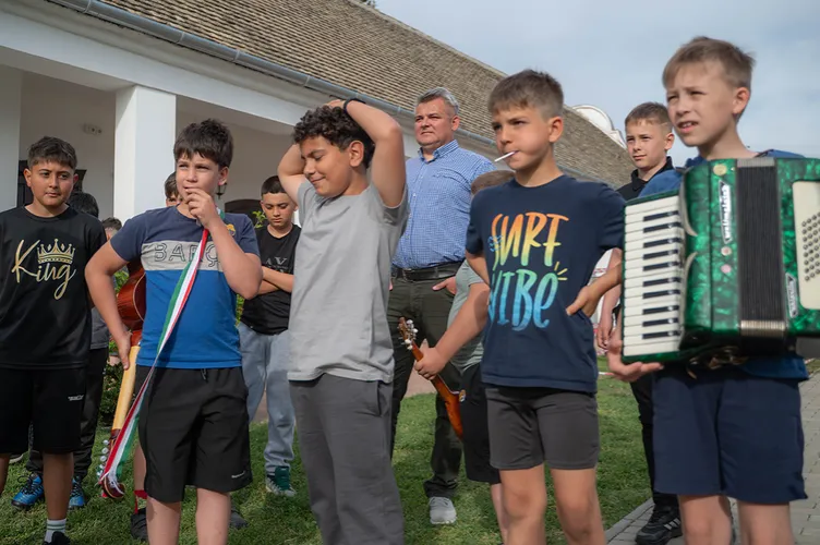 Gyerekek állnak az udvaron, egyikük zöld harmonikát tart, mögöttük több társuk és a zenekarvezető látható.