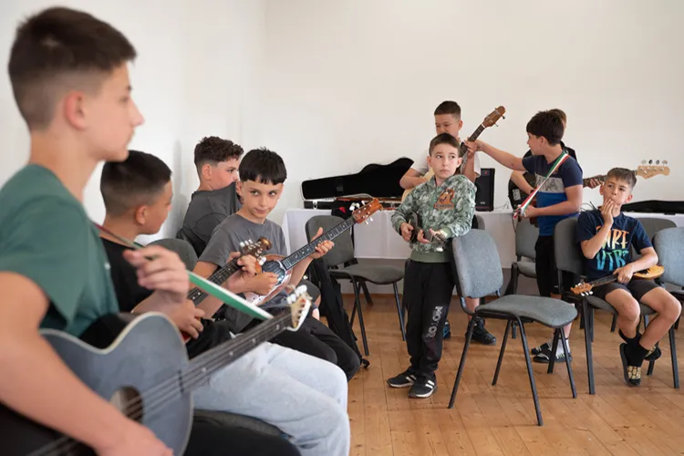 Fiúk ülnek és állnak hangszerekkel a próbateremben, elöl gitár, középen tamburák és üres székek látszanak.