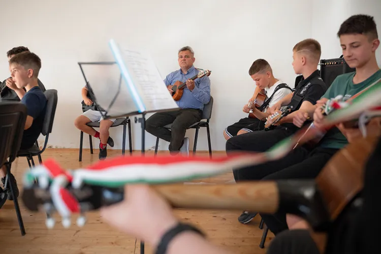 Vezető és a fiúk ülve játszanak gitáron és tamburán, az előtérben kottatartó és egy hangszer látszik.
