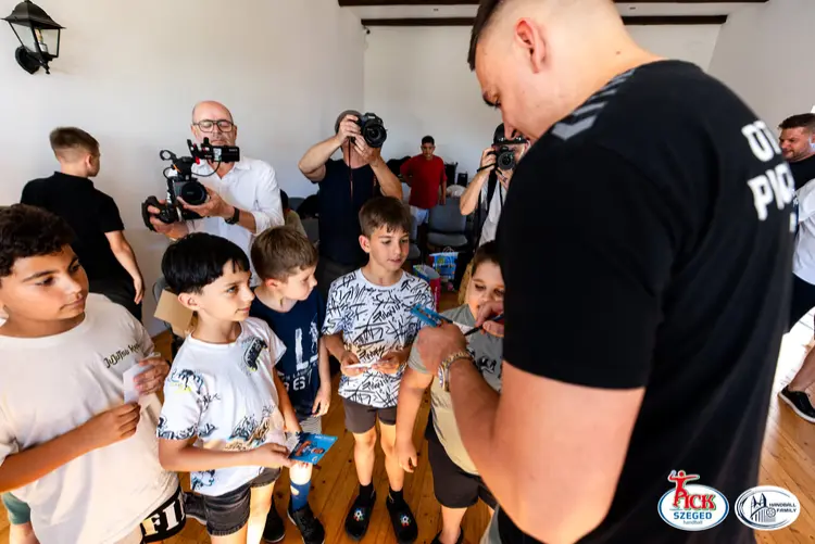 A PICK Szeged játékosa autogramot ad a zenekar gyerektagjainak, miközben a sajtó fotózza a találkozót.