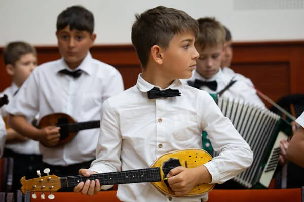 Csokornyakkendős fiú sárga tamburán játszik állva, mögötte harmonikás és tamburás gyerekek zenélnek.