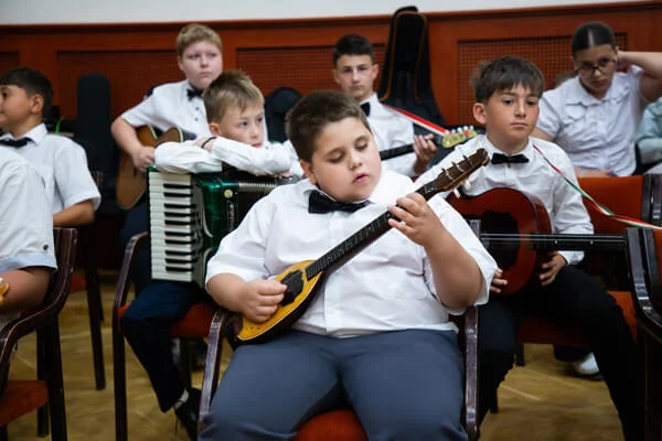 Csokornyakkendős fiú ülve játszik kis tamburán, mögötte harmonikás és gitáros társai figyelnek.