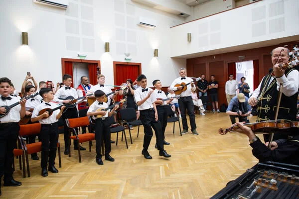 Állva tamburázó fiúk játszanak a felnőtt zenészek felé fordulva, mögöttük egy férfi is penget.