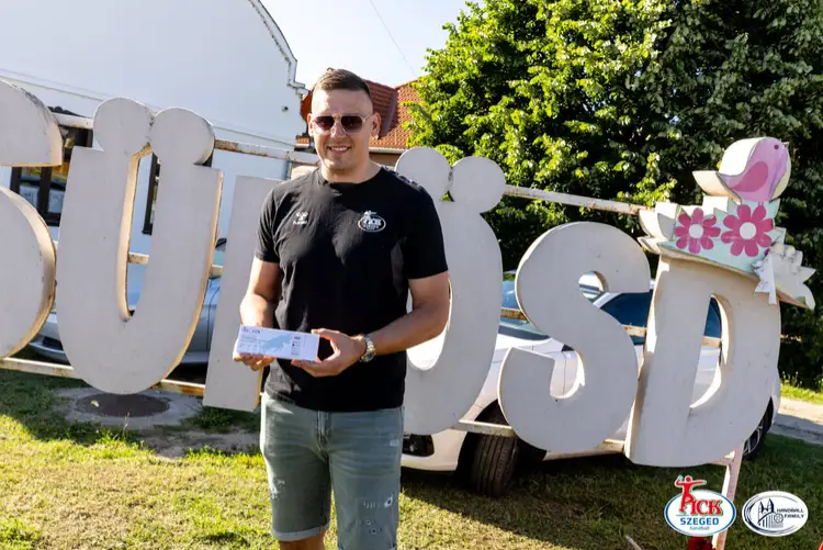 PICK Szeged játékos meccsjegyeket tart a kezében a sükösdi települést jelző felirat előtt