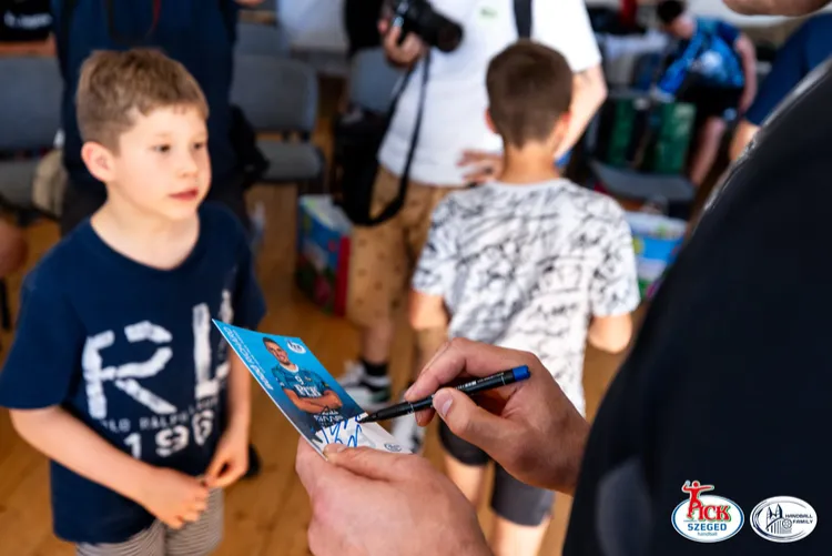 PICK Szeged játékos autogramkártyát ír alá, miközben egy kisfiú várakozik előtte a teremben