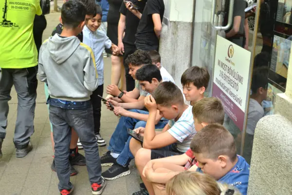 A Sugó Tamburazenekar gyerektagjai egy budapesti üzlet előtt ülve pihennek, többen a telefonjukat nézik.