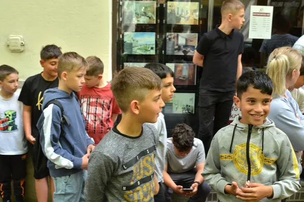 A gyerekek az üzlet előtt várakoznak, az előtérben két fiú beszélget, egyikük a kamerába mosolyog.