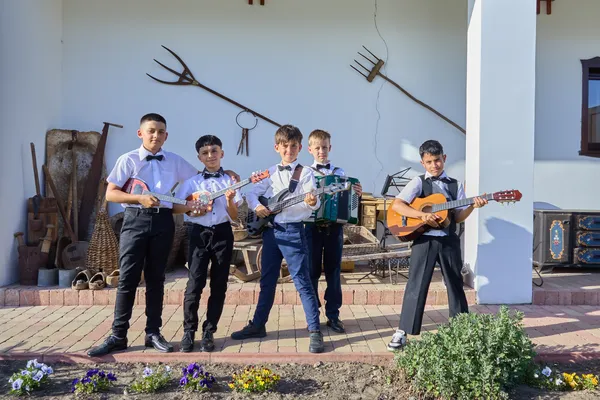 Öt fiú áll hangszerekkel a sükösdi tájház udvarán, tamburát, harmonikát és gitárt tartva.