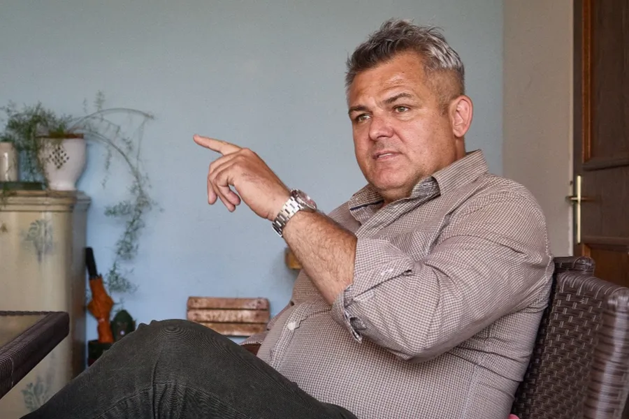 A zenekarvezető ülve beszél és kezével mutat oldalra a sükösdi tájház egyik helyiségében.