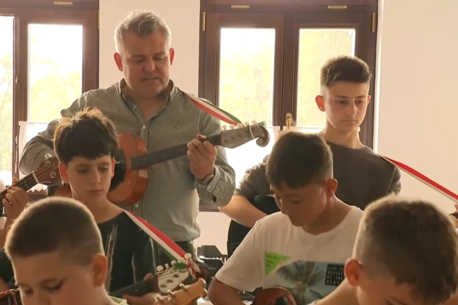 A zenekarvezető tamburán játszik a teremben, miközben körülötte a gyerekek együtt próbálnak.