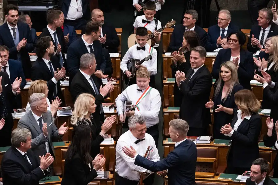 A SUGO Tamburazenekar fiataljai bevonulnak az Országház üléstermébe, miközben a jelenlévők állva tapsolnak.