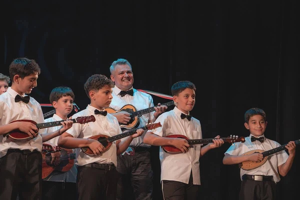 A zenekarvezető mosolyogva tamburázik a gyerekek mögött, miközben a fiúk hangszereikkel állnak a színpadon.