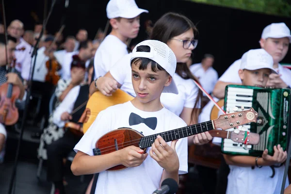Egy fehér sapkás fiatal tamburás közelről a főpróbán, háttérben gitáros és harmonikás zenésztársaival.